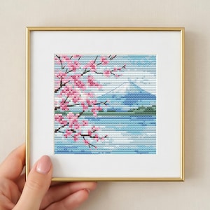 Può includere: Un ricamo a punto croce incorniciato raffigurante un paesaggio giapponese. L'opera presenta una montagna innevata, uno specchio d'acqua e rami di fiori di ciliegio rosa. La cornice è dorata.