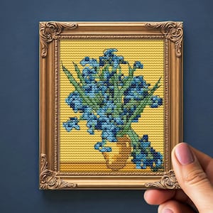 Pode incluir: Uma obra de arte em ponto cruz emoldurada com um buquê de flores azuis em um vaso dourado. A obra de arte está em um fundo amarelo e emoldurada em uma moldura dourada ornamentada. A peça é segurada por uma mão contra um fundo azul escuro.