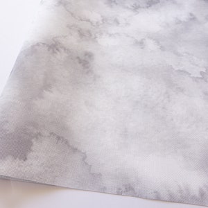 Könnte beinhalten: Nahaufnahme eines Stoffes mit aquarellartigem Design. Der Stoff hat eine weiße Basis mit weichen, grauen wolkenartigen Mustern. Die Textur scheint ein dicht gewebtes Material zu sein, möglicherweise Leinwand oder ein ähnliches Textil.