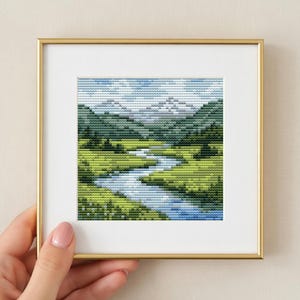 Può includere: Un ricamo a punto croce quadrato che raffigura un paesaggio con un fiume blu tortuoso che scorre attraverso prati verde brillante verso montagne grigie in lontananza sotto un cielo azzurro, racchiuso in una sottile cornice in metallo dorato.