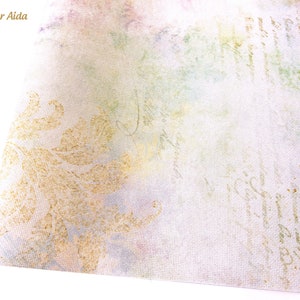 May include: A close-up of a white fabric with a gold glitter pattern. The fabric has a subtle watercolor effect in shades of pink, green, and blue. The text "Glitter Aida" is visible in the top left corner.