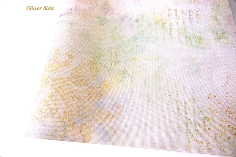 May include: A close-up of a white fabric with a gold glitter pattern. The fabric has a subtle watercolor effect in shades of pink, green, and blue. The text "Glitter Aida" is visible in the top left corner.