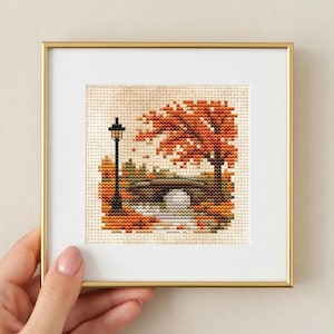May include: A square cross-stitch embroidery in a gold frame depicting an autumn park scene with a black lamppost, a stone bridge over a stream, and trees with orange and brown foliage against a light beige fabric background.