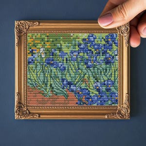 Könnte beinhalten: Ein gerahmtes Kreuzstichbild von Iris in einem goldfarbenen, verzierten Rahmen. Das Kunstwerk zeigt blaue und grüne Iris mit einem braunen und grünen Hintergrund. Der Rahmen wird von einer Hand vor einem dunkelblauen Hintergrund gehalten.