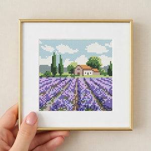 Puede incluir: Un paisaje en punto de cruz enmarcado que representa un campo de lavanda en plena floración, con una pequeña casa y árboles al fondo. El cielo es azul con nubes blancas. El marco es dorado.