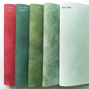 Op de afbeelding: Close-up van vijf stoffen stalen in verschillende kleuren. De stalen zijn gelabeld met nummers en namen: 42-Festive Ruby, 43-Frosted Evergreen, 44-Winter Moss, 45-Snowy Sage en 46-Icy Mint. De stoffen hebben een getextureerde uitstraling.