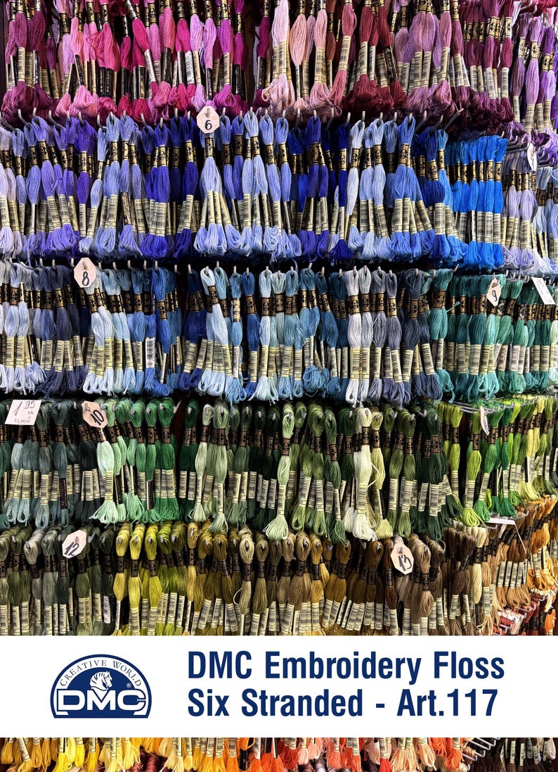 Op de afbeelding: Een muur met DMC borduurgaren, zes draden, Art. 117, in verschillende kleuren. Het garen is gesorteerd op kleur, elke kleur heeft zijn eigen rij. Het garen wordt gepresenteerd op een muur, waarbij elke streng aan een haak hangt.