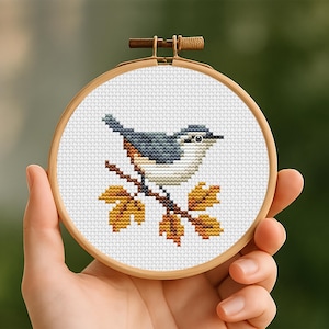 Könnte beinhalten: Ein runder Stickrahmen aus Holz mit einem weißen Stoffstück, das ein Kreuzstichmotiv eines kleinen Vogels mit blauem und weißem Gefieder zeigt, der auf einem braunen Ast mit gelben und orangefarbenen Herbstblättern vor weißem Hintergrund sitzt.