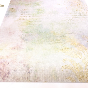May include: A close-up of a piece of white fabric with a subtle, faded floral pattern and a light green, yellow, and pink watercolor wash. The fabric has a textured weave and the text "Glitter Aida" is visible in the top left corner.