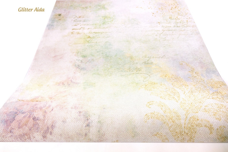 May include: A close-up of a piece of white fabric with a subtle, faded floral pattern and a light green, yellow, and pink watercolor wash. The fabric has a textured weave and the text "Glitter Aida" is visible in the top left corner.