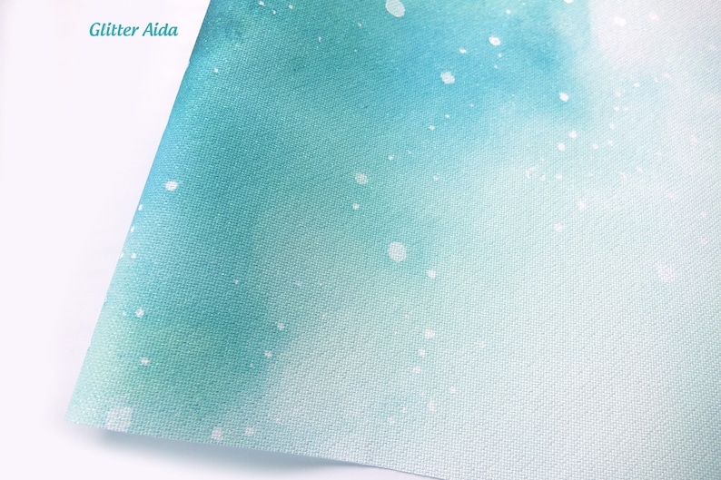 May include: A sheet of Aida fabric featuring a gradient watercolor design that transitions from deep teal to light aqua. The surface is speckled with white glitter accents and includes the text Glitter Aida in the upper left corner.