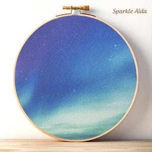 Könnte beinhalten: Ein runder Stickrahmen aus Holz mit einem Stoff, der einen Nachthimmel mit blauen und violetten Nordlichtmustern und verstreuten Sternen zeigt. Der Text Sparkle Aida ist in der oberen rechten Ecke des Bildes aufgedruckt.