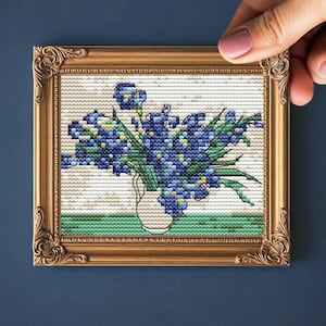 May include: A framed cross-stitch artwork featuring a bouquet of blue flowers in a white vase. The artwork is set against a neutral background and framed in a gold-colored ornate frame. The image is held by a hand.