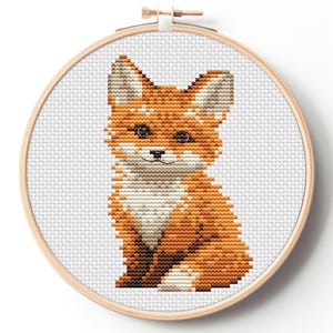 May include: A cross-stitch embroidery hoop depicting a fox. The fox is stitched in orange, brown, and white threads, with detailed work creating a textured look. The embroidery is set against a white background, framed by a wooden hoop.