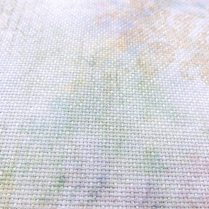 May include: Close-up of a white fabric with a fine weave. The fabric has a subtle, faded pattern of light green, yellow, and brown.