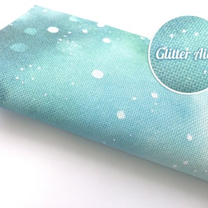 May include: A roll of light blue Aida fabric featuring a white speckled pattern and a subtle glitter finish. A circular inset shows a magnified view of the woven texture and the text Glitter Aida printed in white against the blue fabric background.