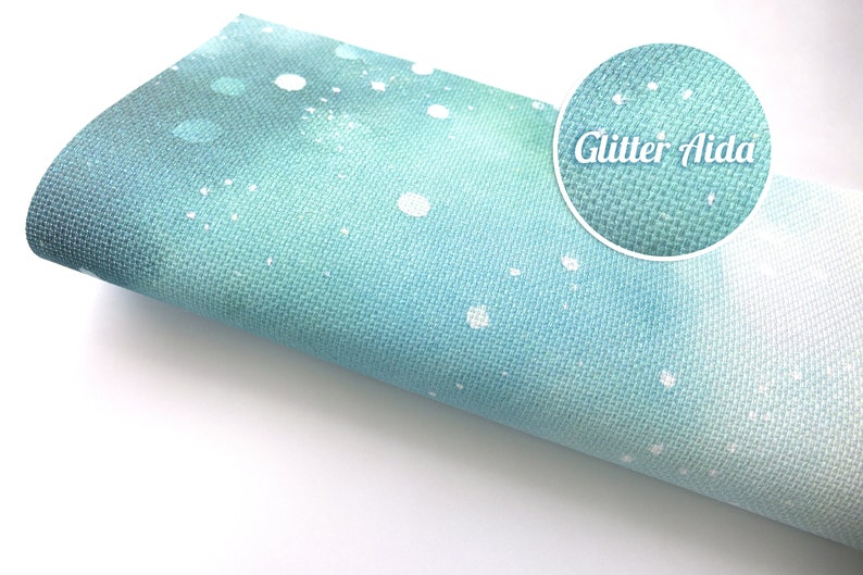 May include: A roll of light blue Aida fabric featuring a white speckled pattern and a subtle glitter finish. A circular inset shows a magnified view of the woven texture and the text Glitter Aida printed in white against the blue fabric background.