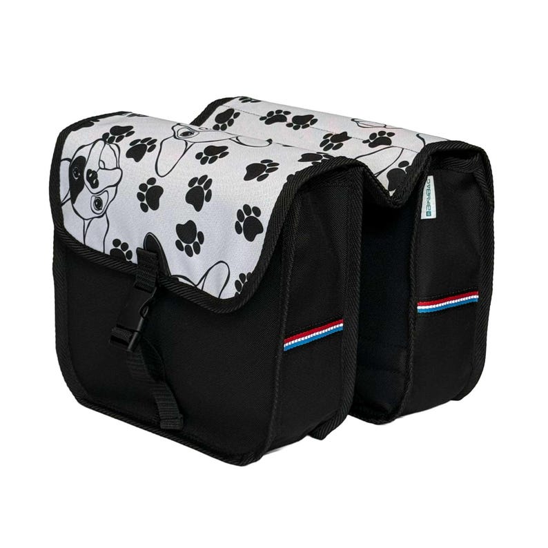 May include: A black double bicycle pannier bag featuring a white flap with a black dog face and paw print pattern. The bag includes a black plastic buckle closure, black trim, and a small blue, white, and red striped accent on the side.