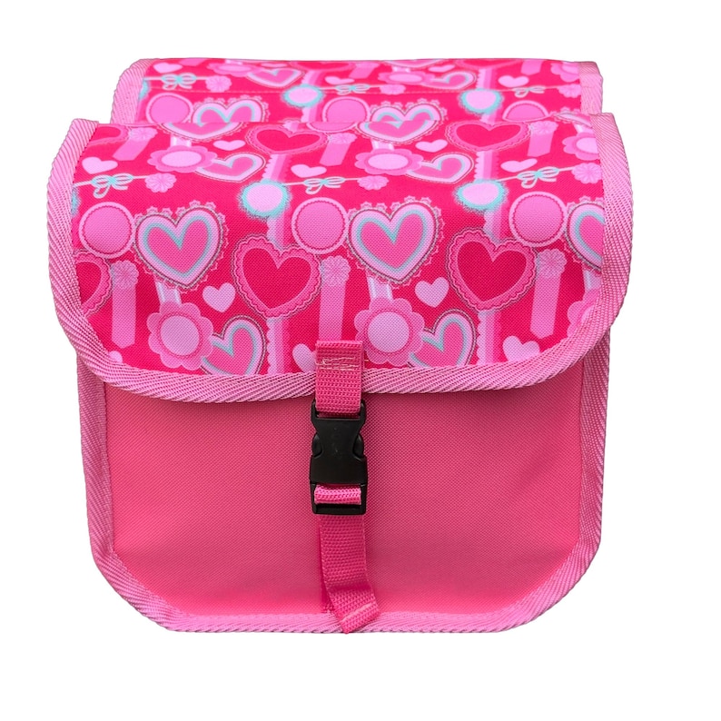 May include: A bright pink bicycle bag featuring a repeating pattern of hearts and circular flower motifs in shades of light pink, white, and cyan. The bag has a flap closure secured with a black plastic side-release buckle and pink trim.