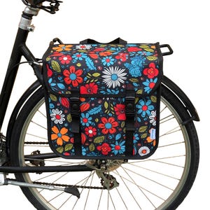 May include: A black bicycle with a floral pannier bag attached to the rear. The bag has a dark background with a colourful array of flowers in red, orange, blue, and white. The bag is secured with black straps and buckles.