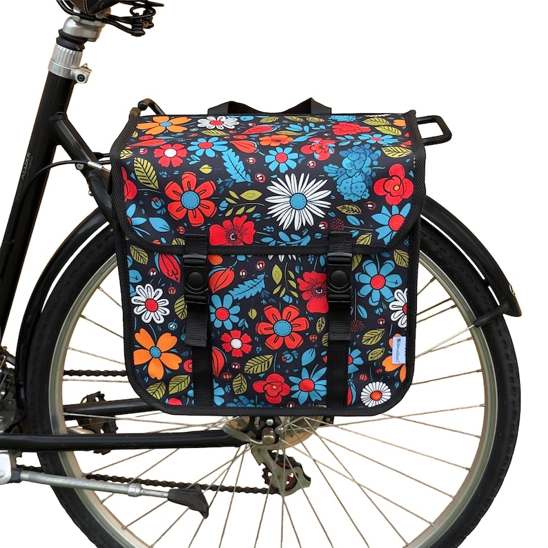 May include: A black bicycle with a floral pannier bag attached to the rear. The bag has a dark background with a colourful array of flowers in red, orange, blue, and white. The bag is secured with black straps and buckles.