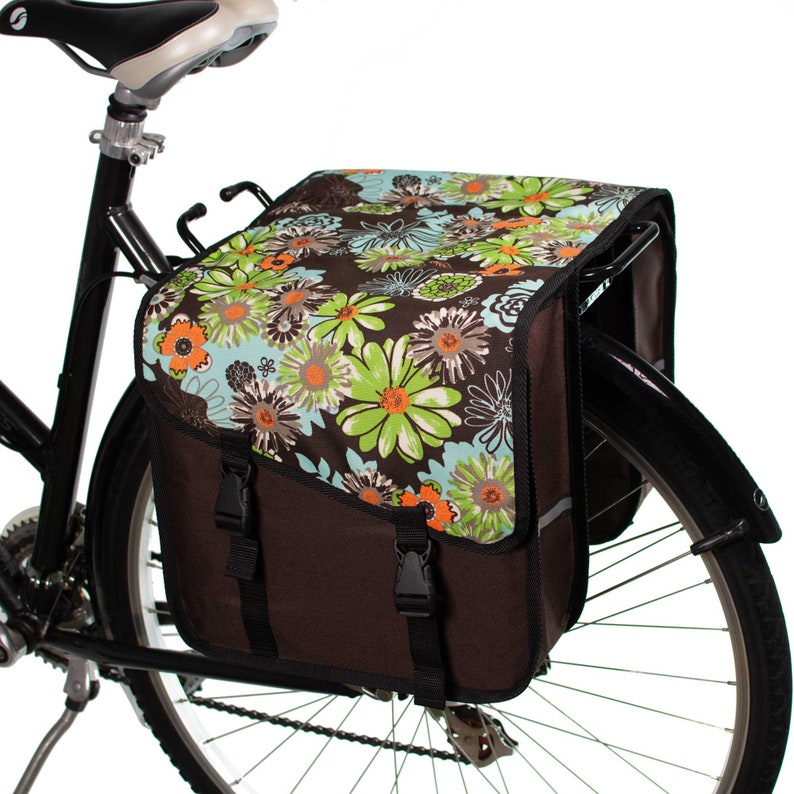 K&ouml;nnte beinhalten: Eine braune Fahrradtasche an einem Gep&auml;cktr&auml;ger mit einer oberen Klappe, die ein mehrfarbiges Blumenmuster in Gr&uuml;n-, Hellblau- und Oranget&ouml;nen aufweist. Die Tasche verf&uuml;gt &uuml;ber zwei schwarze, verstellbare Schnallenriemen am vorderen Verschluss.