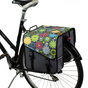 K&ouml;nnte beinhalten: Eine Fahrradtasche, die am Gep&auml;cktr&auml;ger eines schwarzen Fahrrads befestigt ist. Die Tasche hat eine graue Basis mit einem lebendigen Blumenmuster auf der oberen Klappe, einschlie&szlig;lich roter, gelber, blauer und gr&uuml;ner Blumen. Sie hat schwarze Riemen.