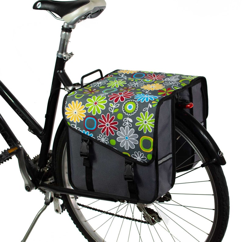 K&ouml;nnte beinhalten: Eine Fahrradtasche, die am Gep&auml;cktr&auml;ger eines schwarzen Fahrrads befestigt ist. Die Tasche hat eine graue Basis mit einem lebendigen Blumenmuster auf der oberen Klappe, einschlie&szlig;lich roter, gelber, blauer und gr&uuml;ner Blumen. Sie hat schwarze Riemen.