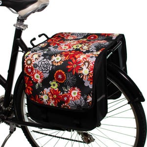 Pu&ograve; includere: Una borsa da bicicletta nera con una stampa floreale con fiori rossi, arancioni e bianchi su sfondo nero. La borsa &egrave; fissata al portapacchi posteriore di una bicicletta.