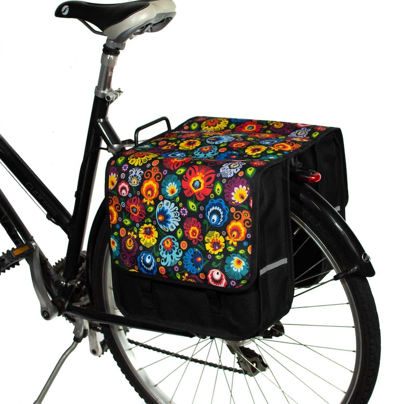 Pu&ograve; includere: Bicicletta nera con una borsa da portapacchi a fantasia floreale fissata al portapacchi posteriore. La borsa da portapacchi ha una base nera e un motivo floreale colorato con fiori rossi, gialli, blu, verdi e bianchi.