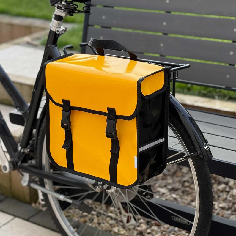 May include: A bright yellow bicycle pannier bag mounted on a black bicycle. The bag features a flap closure with two black buckle straps. It has black trim and a reflective strip. The bag is designed for carrying items.