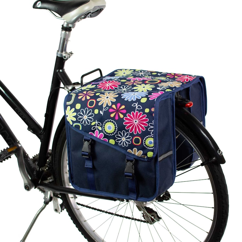 K&ouml;nnte beinhalten: Eine marineblaue Fahrradtasche, die am Gep&auml;cktr&auml;ger eines schwarzen Fahrrads befestigt ist. Die Tasche hat eine Klappe mit einem bunten Blumenmuster in Pink, Gelb, Hellblau und Wei&szlig;, gesichert durch zwei schwarze Kunststoffschnallen am marineblauen Korpus.