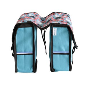 May include: A double bicycle pannier bag in bright blue with black trim and a top flap featuring a repeating pattern of stylised cats in red, white, and black. The bag includes side buckles, reflective horizontal strips, and a small white brand tag.
