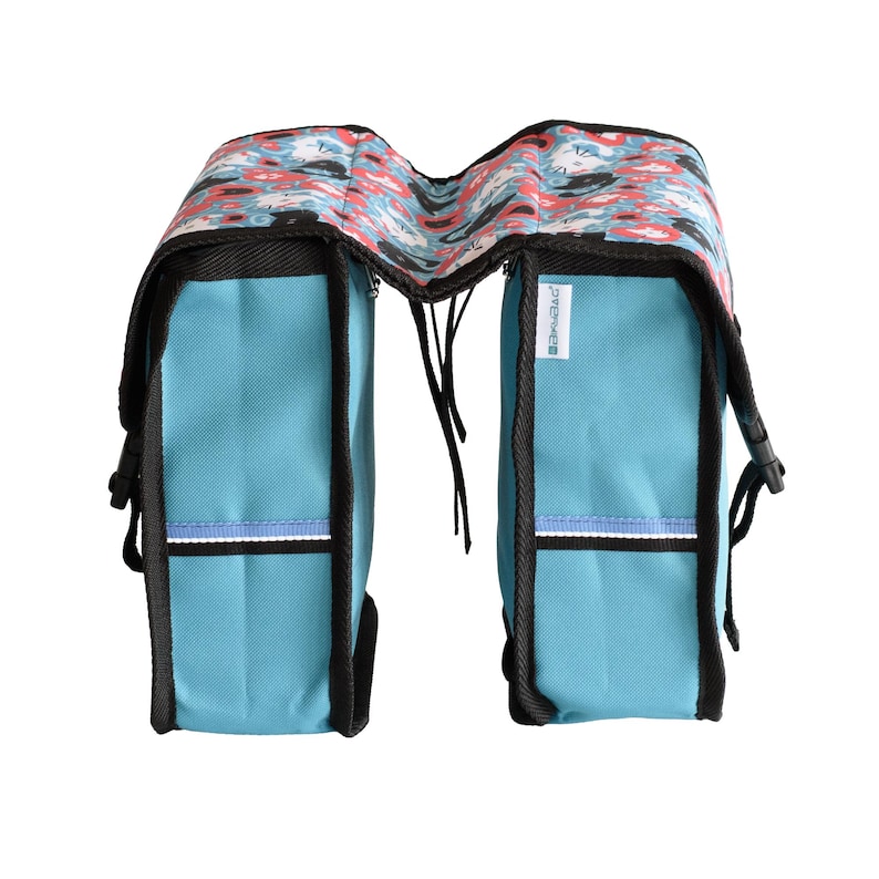 May include: A double bicycle pannier bag in bright blue with black trim and a top flap featuring a repeating pattern of stylised cats in red, white, and black. The bag includes side buckles, reflective horizontal strips, and a small white brand tag.