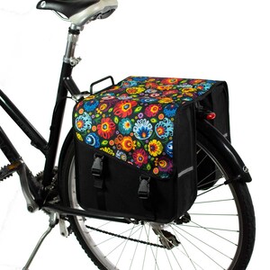 K&ouml;nnte beinhalten: Eine schwarze Fahrradtasche, die am Gep&auml;cktr&auml;ger eines Fahrrads befestigt ist. Die Tasche hat ein lebhaftes, mehrfarbiges Blumenmuster auf der oberen Klappe, die mit zwei schwarzen Kunststoffschnallen &uuml;ber einem schwarzen Hauptfach gesichert ist.