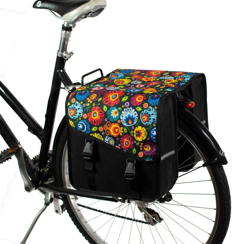 K&ouml;nnte beinhalten: Eine schwarze Fahrradtasche, die am Gep&auml;cktr&auml;ger eines Fahrrads befestigt ist. Die Tasche hat ein lebhaftes, mehrfarbiges Blumenmuster auf der oberen Klappe, die mit zwei schwarzen Kunststoffschnallen &uuml;ber einem schwarzen Hauptfach gesichert ist.