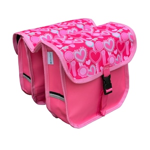May include: A pair of bright pink bicycle pannier bags featuring a repeating pattern of hearts and circular floral motifs on the top flaps. Each bag has a black plastic buckle closure, pink fabric trim, and a small reflective strip on the side.