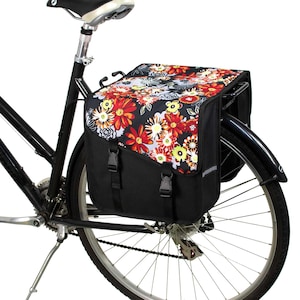 K&ouml;nnte beinhalten: Eine schwarze Fahrradtasche, die am Gep&auml;cktr&auml;ger eines Fahrrads befestigt ist. Die Tasche verf&uuml;gt &uuml;ber eine Klappe mit einem lebendigen Blumenmuster in Rot-, Orange-, Gelb- und Wei&szlig;t&ouml;nen auf schwarzem Grund, gesichert mit zwei schwarzen Kunststoffschnallen.