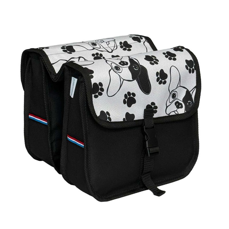 May include: A black double bicycle pannier bag featuring a white flap with a repeating pattern of black and white dog faces and paw prints. The bag includes a black plastic buckle closure and a small blue, white, and red striped fabric tag.