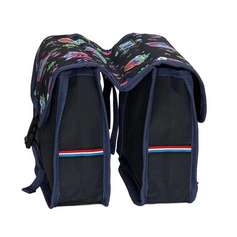 May include: A black double bicycle pannier bag featuring a colourful owl pattern on the top flaps. The bag has dark blue trim, side buckles, and a horizontal stripe detail in red, white, and light blue on the front of each compartment.