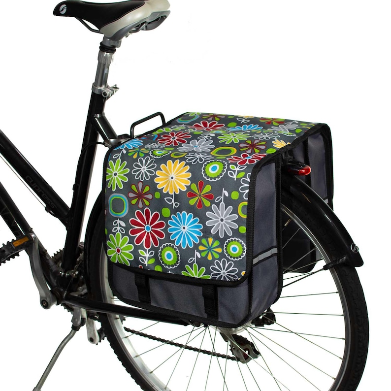 Pu&ograve; includere: Una bicicletta nera con una borsa a pannier con motivo floreale fissata al portapacchi posteriore. La borsa ha una base grigia e un design floreale colorato con fiori rossi, gialli, blu e verdi.