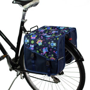 K&ouml;nnte beinhalten: Eine dunkelblaue Fahrradtasche, die am Gep&auml;cktr&auml;ger eines schwarzen Fahrrads befestigt ist. Die Tasche hat eine Klappe mit einem bunten Eulen- und Blumenmuster in Rosa, Hellblau und Limettengr&uuml;n, die mit zwei schwarzen Kunststoffschnallen gesichert ist.