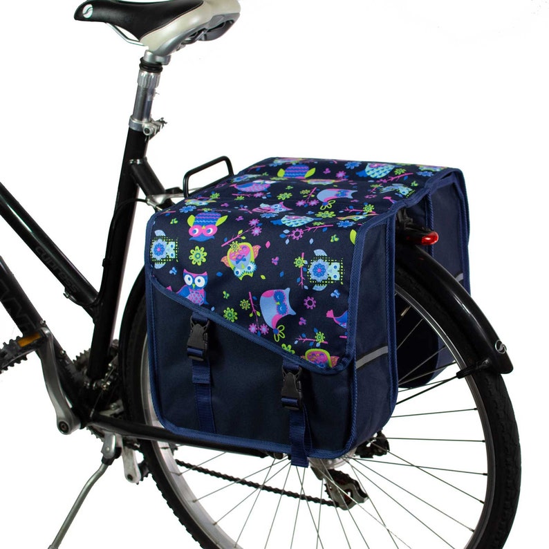 K&ouml;nnte beinhalten: Eine dunkelblaue Fahrradtasche, die am Gep&auml;cktr&auml;ger eines schwarzen Fahrrads befestigt ist. Die Tasche hat eine Klappe mit einem bunten Eulen- und Blumenmuster in Rosa, Hellblau und Limettengr&uuml;n, die mit zwei schwarzen Kunststoffschnallen gesichert ist.