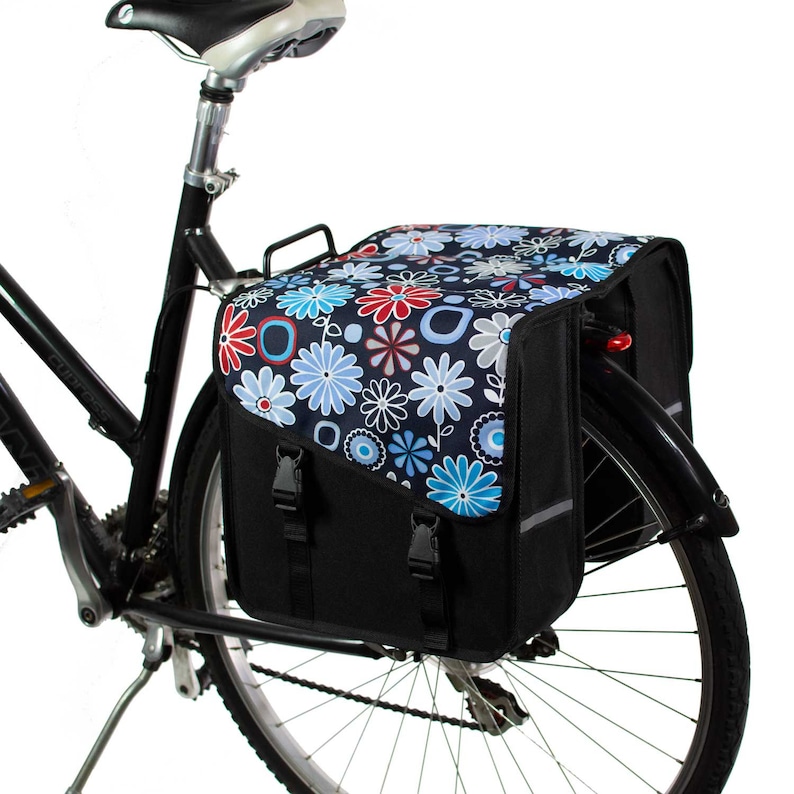 K&ouml;nnte beinhalten: Eine schwarze Fahrradtasche, die am Gep&auml;cktr&auml;ger eines Fahrrads befestigt ist. Die Tasche hat eine obere Klappe mit dunkelblauem Hintergrund und einem wiederkehrenden Muster aus hellblauen, roten und wei&szlig;en Blumen und geometrischen Formen.