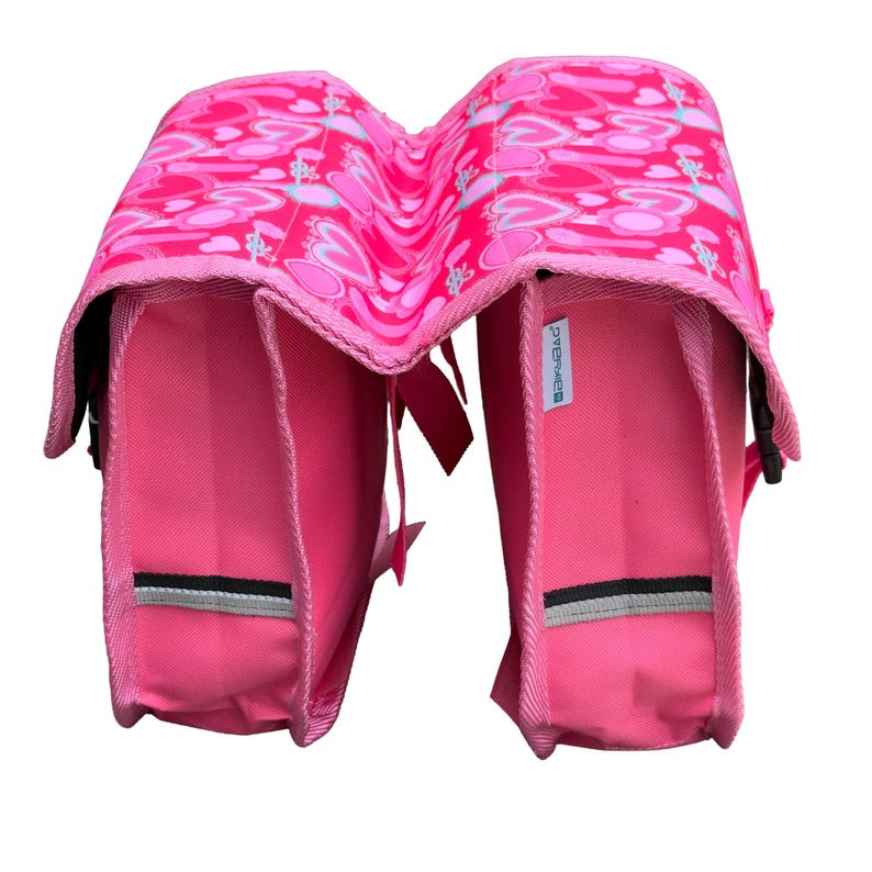 May include: A bright pink double bicycle pannier bag featuring a repeating pattern of hearts and arrows on the top flaps. The bag has solid pink side panels, black and grey reflective strips, and a small white tag with the text "BASIL" on the side.