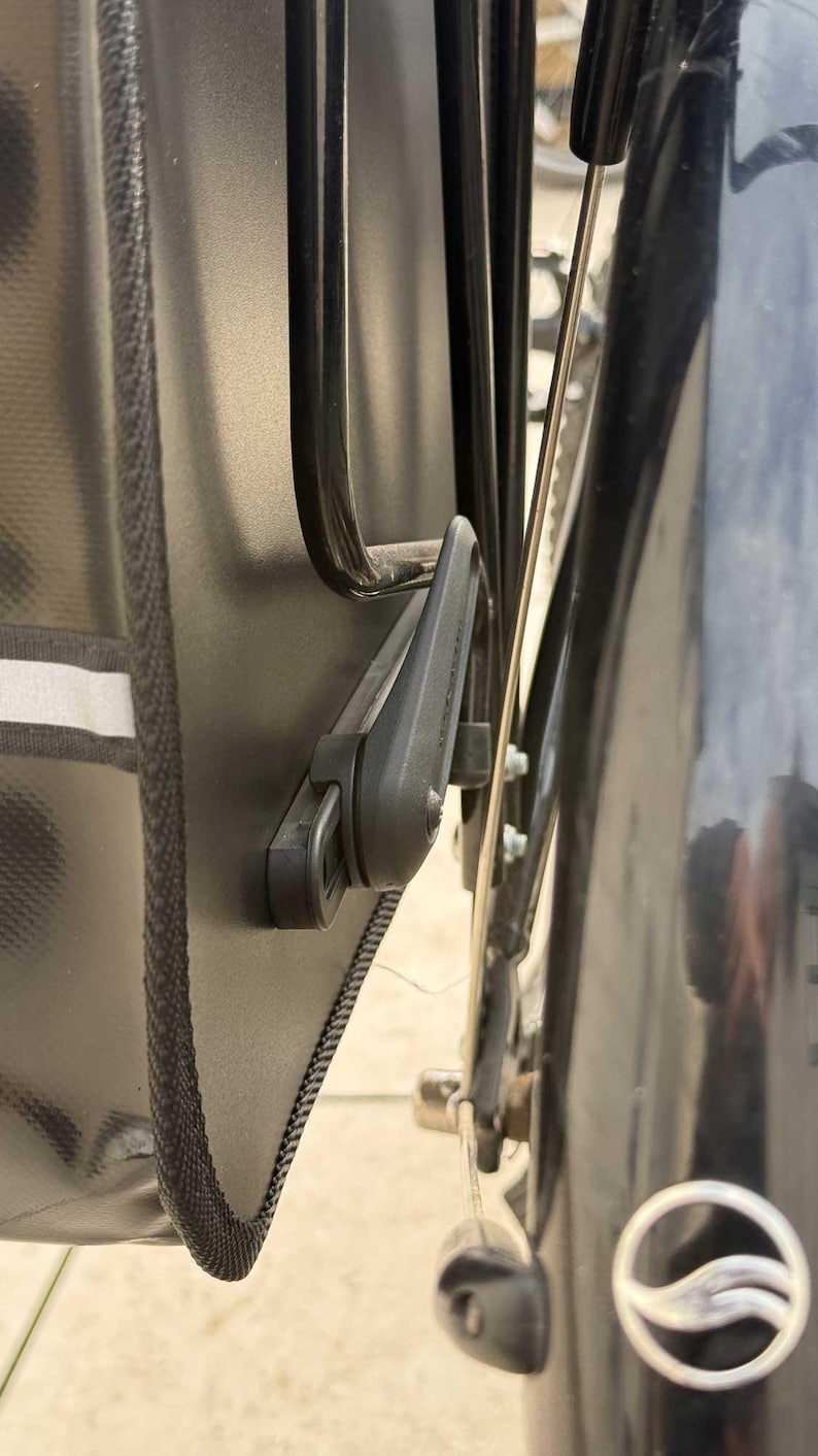 May include: Close-up of a black bicycle pannier bag attached to a bike rack. The bag has a reflective stripe and a black locking mechanism. The bike's black mudguard and wheel are also visible.