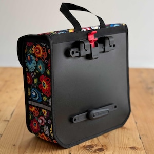 May include: Black bicycle pannier bag with a vibrant floral design. The bag has a black handle, red and black clips, and a reflective strip. The floral pattern features red, blue, yellow, and green flowers on a black background.