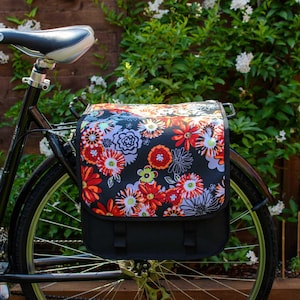Pu&ograve; includere: Una bicicletta nera con una borsa da portapacchi a fantasia floreale fissata al portapacchi posteriore. La borsa &egrave; nera con un design floreale colorato e ha una patta nera.