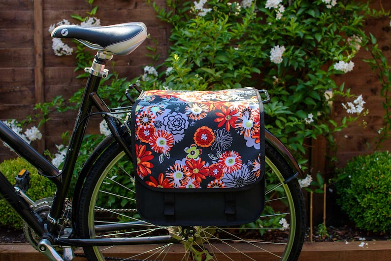 Pu&ograve; includere: Una bicicletta nera con una borsa da portapacchi a fantasia floreale fissata al portapacchi posteriore. La borsa &egrave; nera con un design floreale colorato e ha una patta nera.
