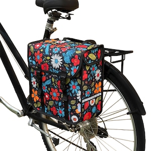 Puede incluir: Alforja de bicicleta negra con un colorido estampado floral de flores rojas, azules, naranjas y blancas. La bolsa está sujeta al portaequipajes trasero de una bicicleta. Tiene una banda reflectante y dos hebillas negras.